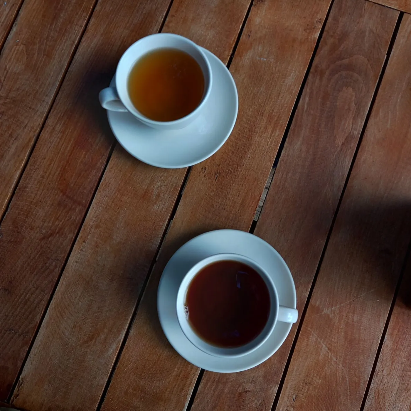 Two cups of black tea on white saucers, set across from each other on a warm-toned wooden table, seen from above