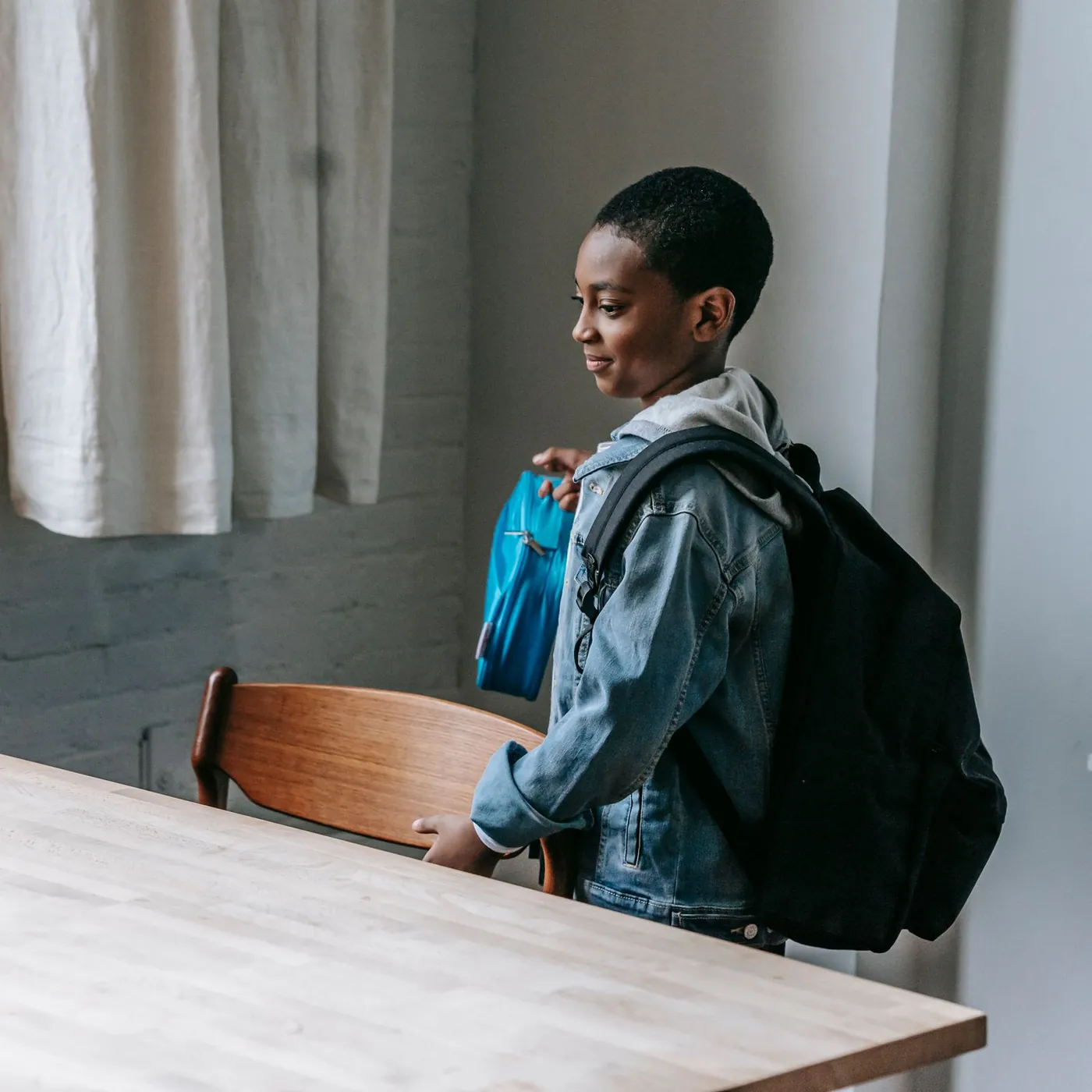 A boy in a denim jacket and backpack, lunch bag in hand, pulling out a wooden chair at a classroom table by a curtained window