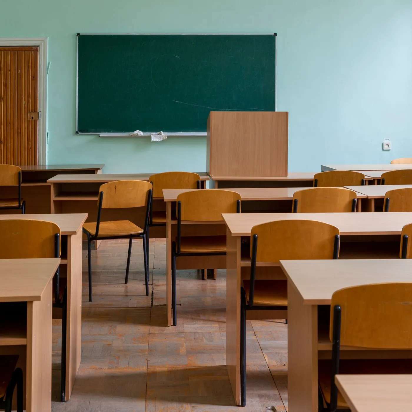 An empty classroom of light wooden desks and chairs facing a green chalkboard and lectern, with a panelled door and a bright window