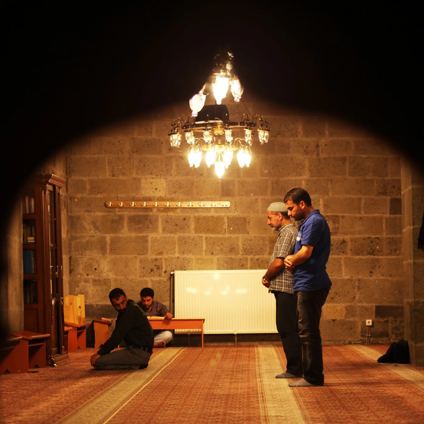 Men praying and reading in a stone-walled masjid under a glowing chandelier, framed by a dark arch