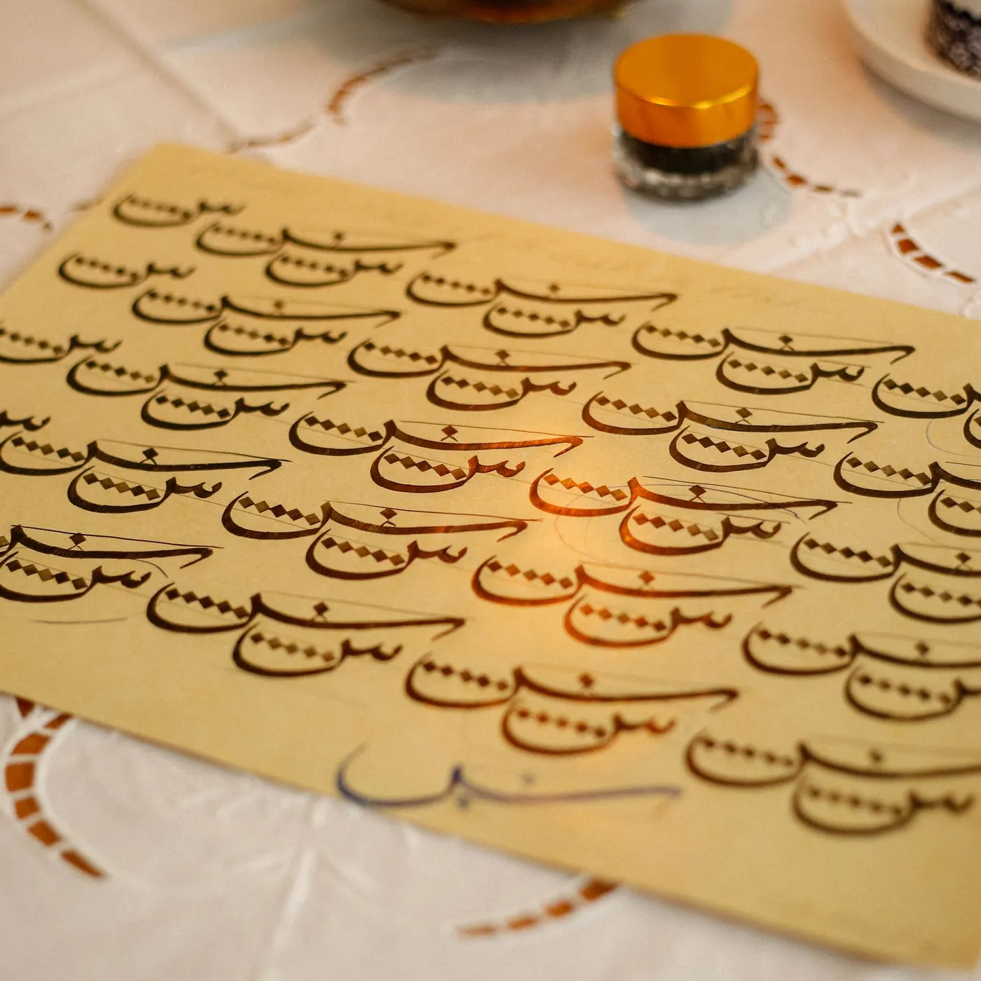 A calligraphy practice sheet covered in repeated Arabic letter forms in brown ink, beside reed pens and a small ink pot