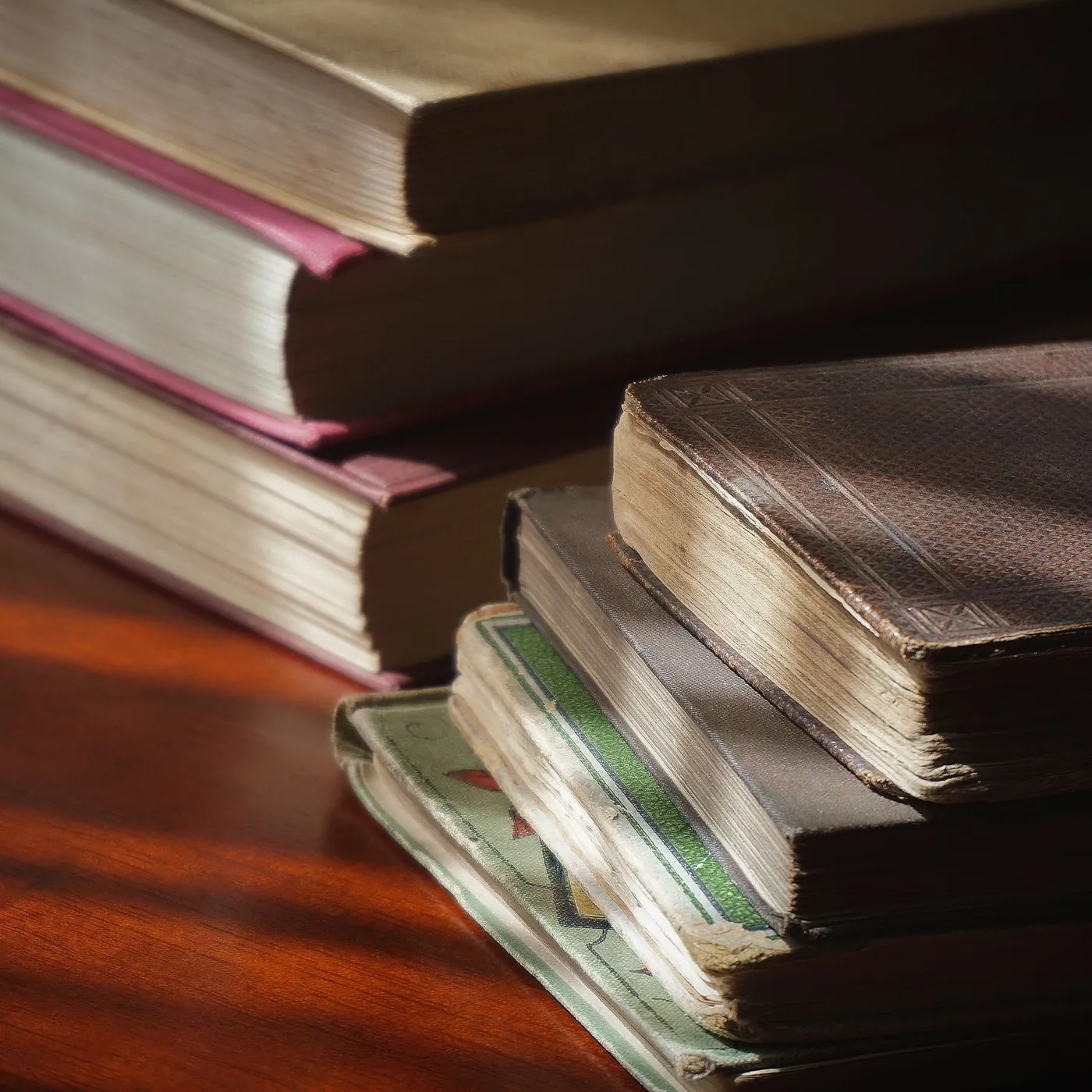 Stacks of old cloth-bound books on a polished wooden table, striped with afternoon window light