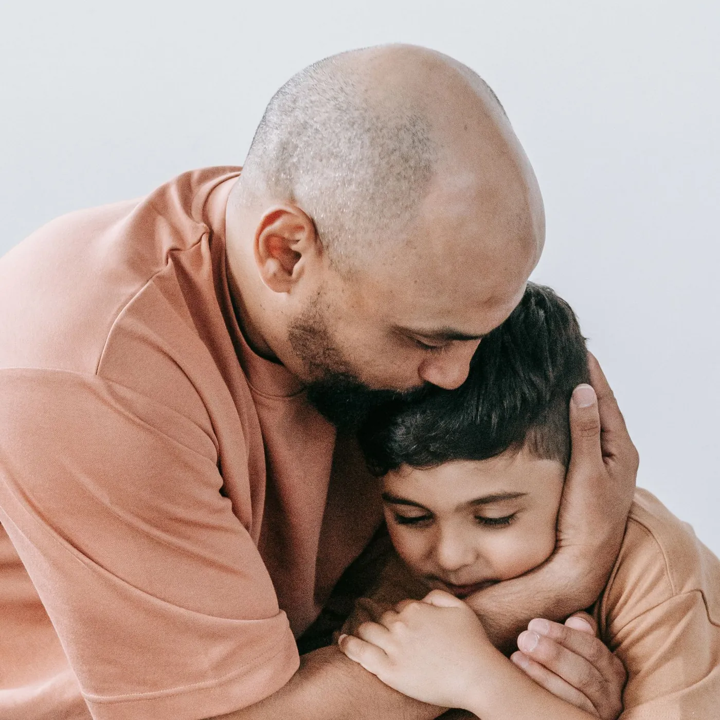 A father in a peach T-shirt holding his young son close, kissing the top of his head as the boy rests his cheek in his hand