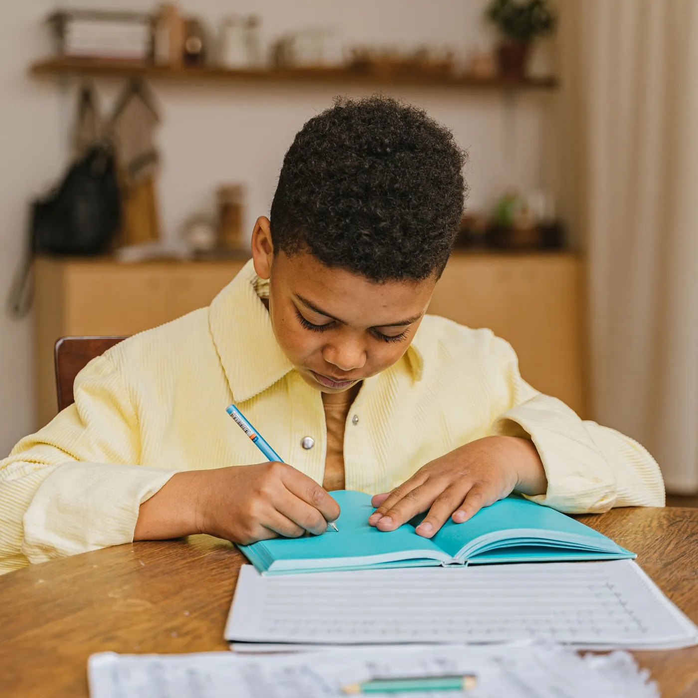 A young boy in a pale yellow shirt bends over a turquoise exercise book, carefully writing at a wooden table