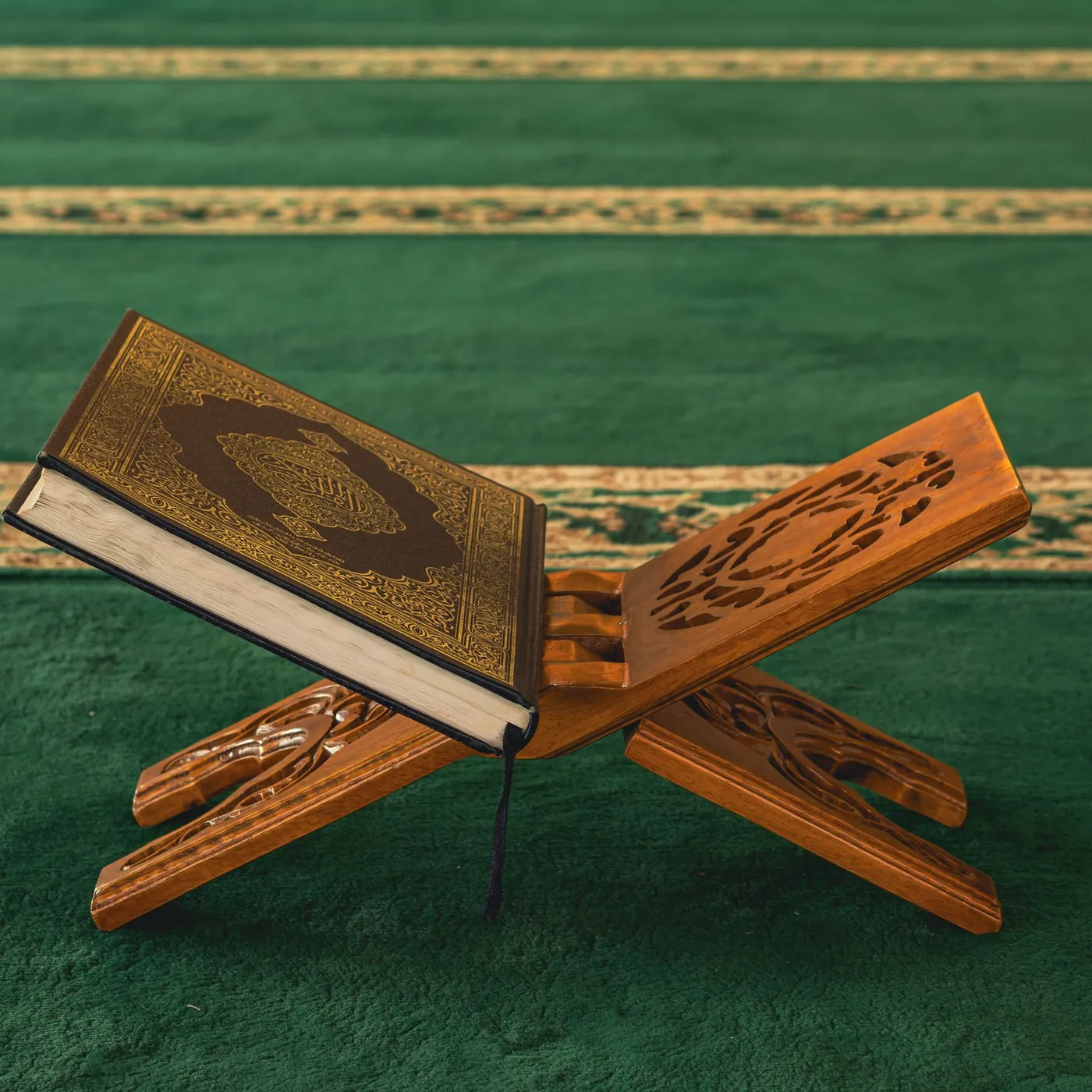 A closed Qur’an with a gold-tooled brown cover resting on a carved wooden rehal on green mosque carpet