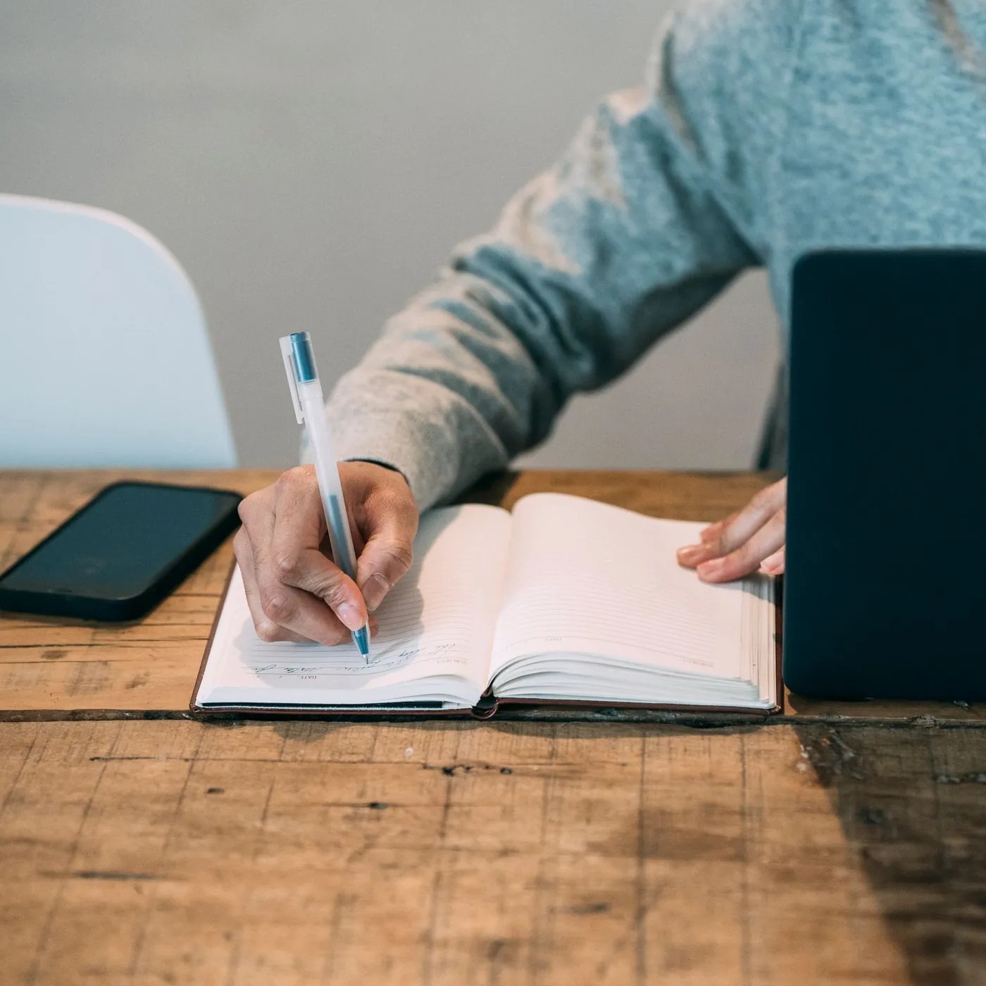 A student writes plans in a lined planner on a rustic wooden table, a phone to one side and a closed dark laptop beside him