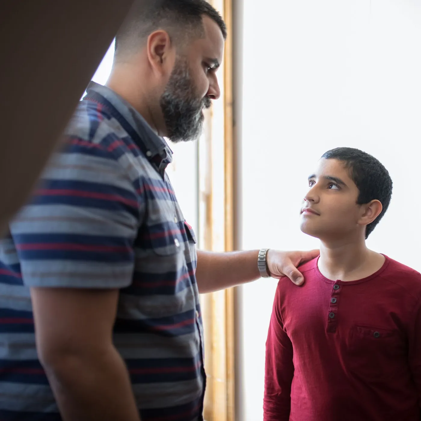 A bearded father rests a steady hand on his teenage son's shoulder as the boy looks up at him beside a bright window