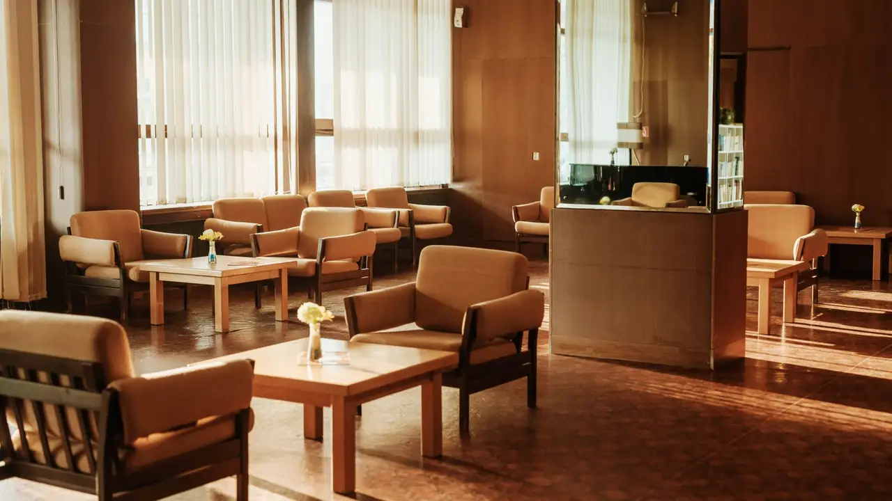 An empty waiting lounge in low amber sunlight: tan armchairs around blond-wood tables, small vases of flowers, and a glazed staff window at the far side.