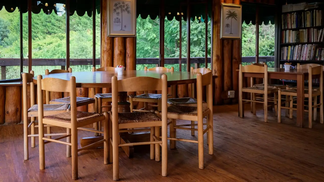 A timber-framed study room with peeled log posts, rush-seat chairs pulled round two tables, and tall windows onto green forest.