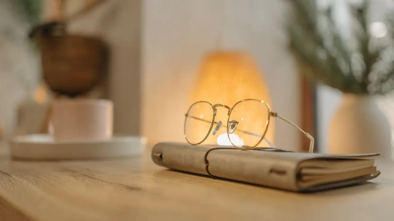 A tan leather journal with gold-rimmed reading glasses resting on it, on an oak tabletop under a warm shaded lamp.