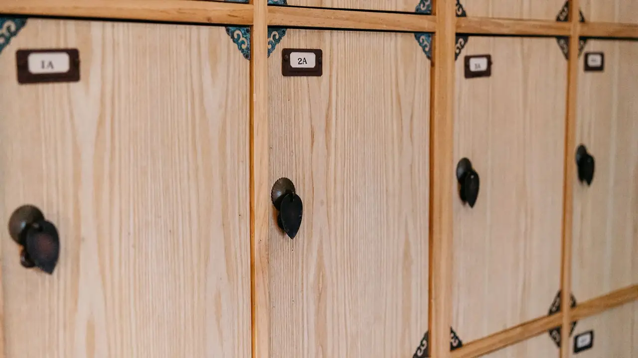 Blond pine coat-room lockers photographed close, the doors labeled 1A, 2A and 3A in small brass card holders with leaf-shaped iron pulls.