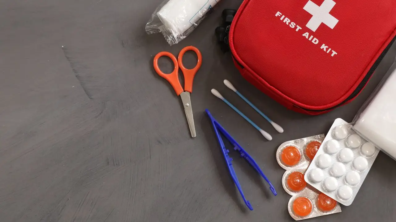 A red first aid kit pouch on mid-dark slate, with orange-handled scissors, tweezers, swabs and gauze laid out beside it.