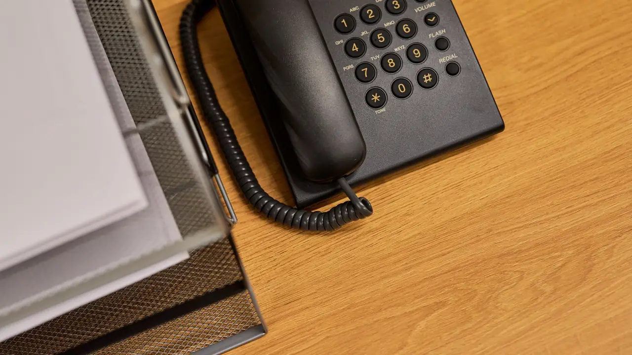An unbranded black desk telephone on honey oak, with a wire paper tray alongside.